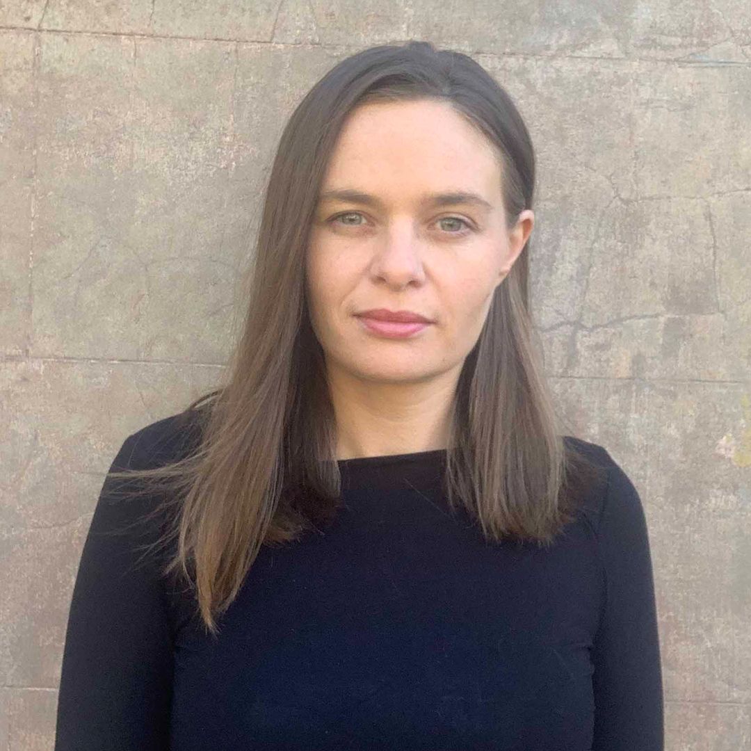 Photo of a women with brown hair standing against a stone wall staring into camera wearing black shirt. 